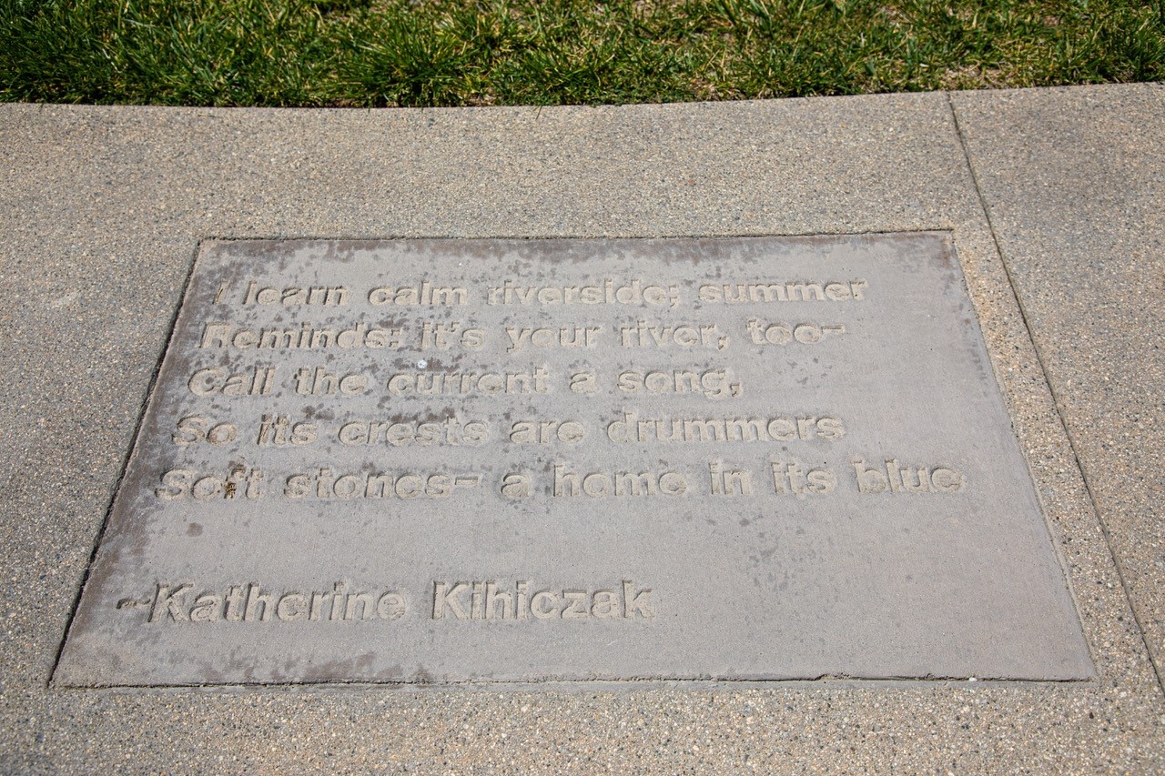 Sidewalk Poetry. Poem reads "I learn calm riverside; summer reminds: it's your river too--call the current a song, so its crests are drummers, soft stones--a home in its blue." - Katherine Kihiczak