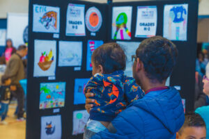 Parent holding their small child as they look through the Youth Arts exhibition. The background shows black walls lined with various youth art entries.