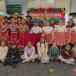 Children from the Ballet Folklórico dressed in traditional Ballet Folklórico dresses standing while more children are sitting in front of them.