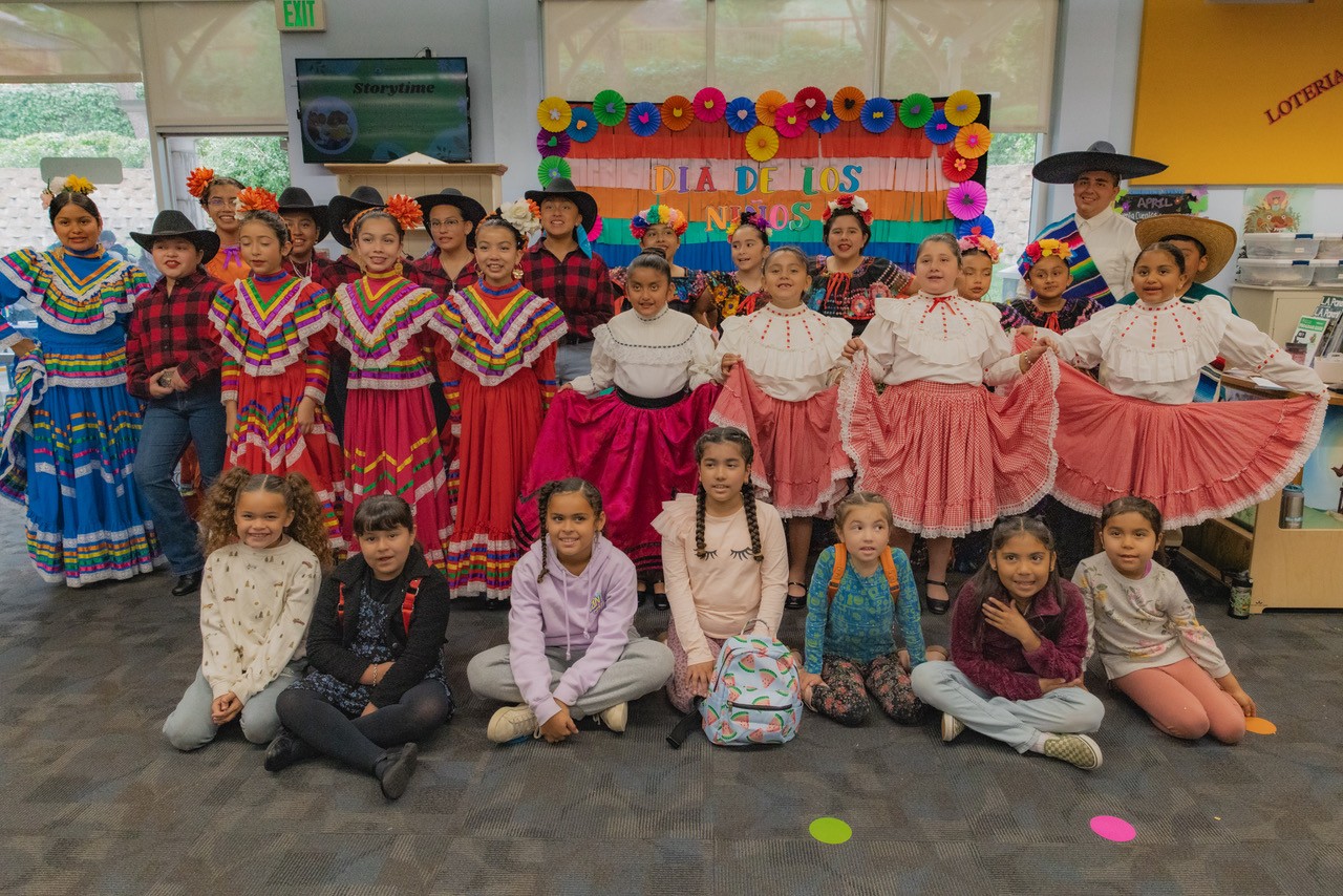 Children from the Ballet Folklórico dressed in traditional Ballet Folklórico dresses standing while more children are sitting in front of them.